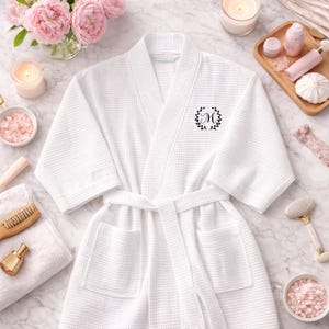 Może przedstawiać: Biały szlafrok o strukturze wafla z czarnym monogramem "M" w wieńcu. Szlafrok ma pas i kieszenie. Obraz zawiera akcesoria spa, takie jak świece, kwiaty i produkty do kąpieli, tworząc relaksującą atmosferę.
