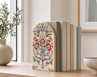 Puede incluir: Un sujetalibros de madera en forma de arco con un panel frontal de color crema que presenta un patrón floral simétrico en tonos rojos, naranjas, azules y verdes. El sujetalibros sostiene una fila de cinco libros de colores sólidos en tonos apagados de gris, beige y verde.
