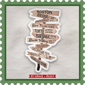 Puede incluir: Una pegatina con señales de madera que apuntan a varias ciudades, como Boston, Seúl y Tokio. Las distancias se muestran en millas y kilómetros. La pegatina tiene un borde blanco y el texto "BY GRACE + ROCKY" en la parte inferior.
