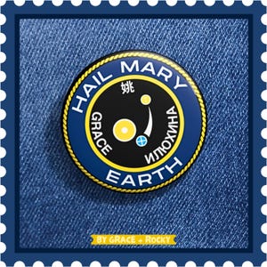 Puede incluir: Un pin redondo, azul marino y negro, con borde dorado y las palabras "HAIL MARY EARTH" en blanco. El pin también presenta las palabras "GRACE" e "ИЛЮХИНА" y un pequeño gráfico. El pin está sobre un fondo de mezclilla.