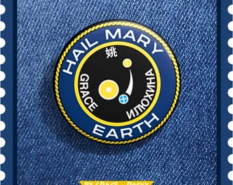 May include: A round, navy blue and black pin with gold trim and the words "HAIL MARY EARTH" in white. The pin also features the words "GRACE" and "ИЛЮХИНА" and a small graphic. The pin is on a denim background.