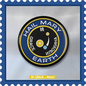 Könnte beinhalten: Ein blauer und gelber Knopf mit dem Text "Hail Mary Earth" und einer Grafik eines Planeten mit einer Mondsichel. Der Text "Grace" ist vertikal auf der linken Seite der Grafik geschrieben und der Text "Илюхина" ist vertikal auf der rechten Seite der Grafik geschrieben. Der Text "By Grace + Rocky" ist unter dem Knopf geschrieben.