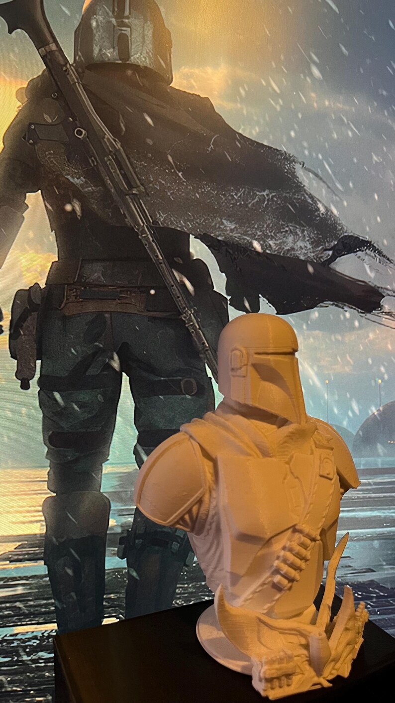 May include: A 3D-printed bust of a Mandalorian character, featuring a helmet and armor details. The bust is light-colored and stands on a black surface, with a background image of the character in a snowy landscape.