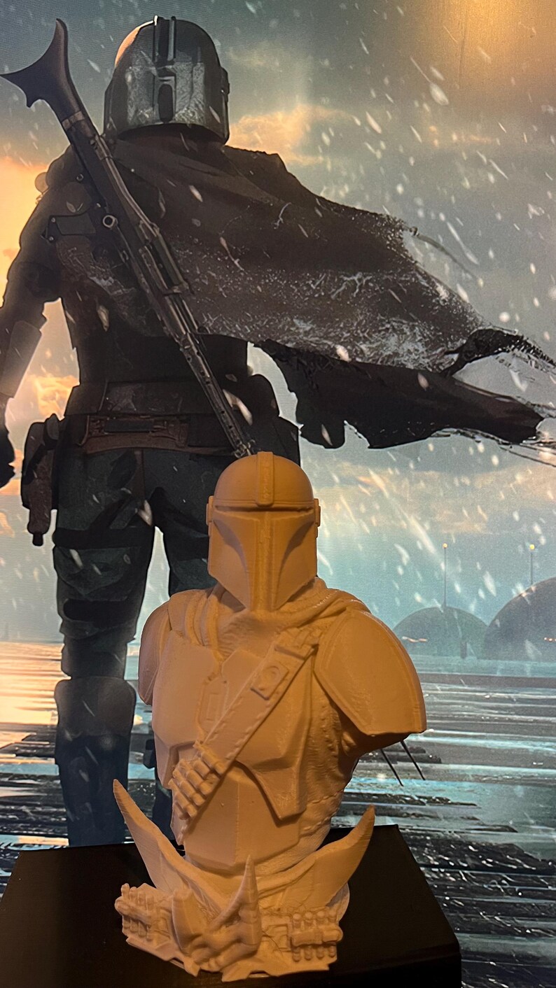 May include: A 3D-printed bust of a Mandalorian character, featuring a helmet and armor. The bust is light beige, with details of the armor visible. A larger image of the character is in the background, with a snowy landscape.