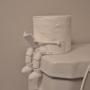 Pode incluir: Um rolo de papel higiênico branco com um suporte decorativo, em forma de pessoa sentada lendo um livro. O rolo de papel higiênico está em cima de uma sanita branca.
