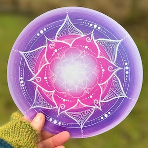 Könnte beinhalten: Ein rundes Mandala-Design in Lila und Rosa mit weißen Akzenten. Das Mandala zeigt ein zentrales blütenartiges Muster mit komplizierten Details und einem Farbverlauf. Das Kunstwerk wird in einer Hand mit einem grünen Handschuh gehalten.