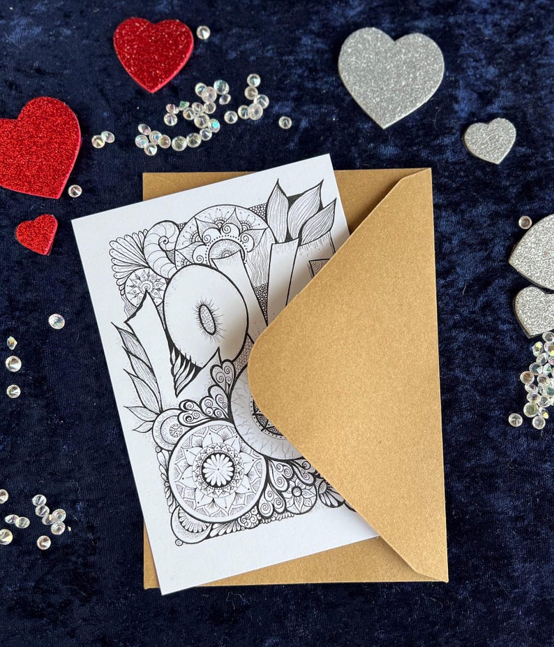 Handgezeichnete Love Postkarte zum Ausmalen mit Mandala- und floralen Mustern und braunem Briefumschlag auf dunklem Stoff dekoriert mit Herz Deko