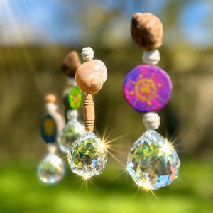 Könnte beinhalten: Ein dekorativer Sonnenfänger mit mehreren Strängen. Jeder Strang hat einen facettierten Kristallkugel, Holzperlen, Muscheln und bemalte Holzscheiben in Lila, Grün und Braun. Der Sonnenfänger ist zum Aufhängen konzipiert.