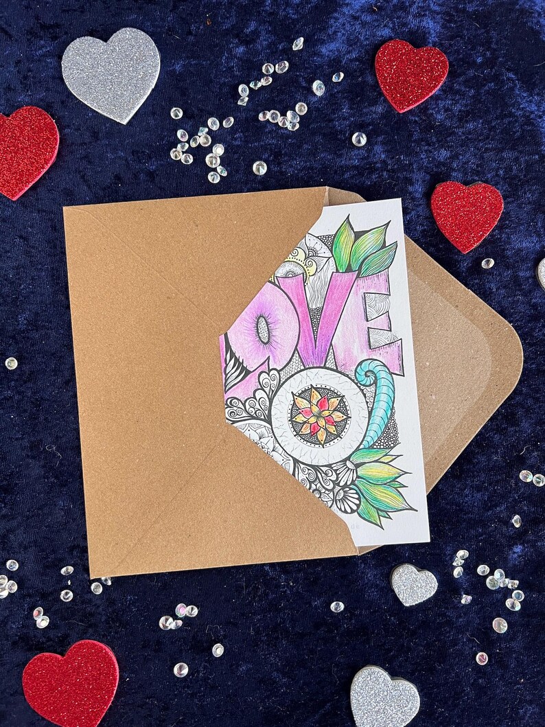 Handgezeichnete Love Postkarte zum Ausmalen (mit Buntstiften angemalt) und braunem Briefumschlag auf dunklem Stoff dekoriert mit Herz Deko