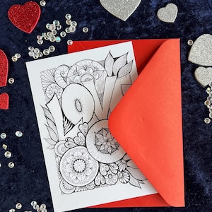 Handgezeichnete Love Postkarte zum Ausmalen mit Mandala- und floralen Mustern in einem roten Briefumschlag liegend auf dunklem Stoff dekoriert mit Herzdeko
