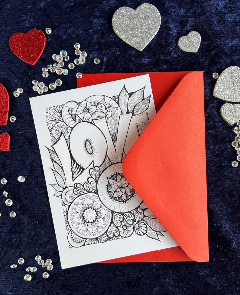 Handgezeichnete Love Postkarte zum Ausmalen mit Mandala- und floralen Mustern in einem roten Briefumschlag liegend auf dunklem Stoff dekoriert mit Herzdeko
