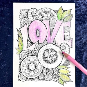 May include: A white card with the word "LOVE" in pink, surrounded by intricate black and white mandala-style designs. Green and yellow leaves accent the design. A pink colored pencil rests on the card.