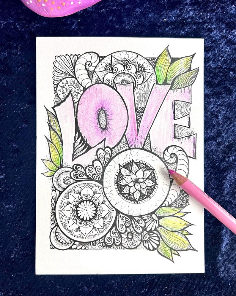 May include: A white card with the word "LOVE" in pink, surrounded by intricate black and white mandala-style designs. Green and yellow leaves accent the design. A pink colored pencil rests on the card.