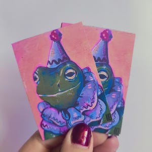 May include: Two rectangular stickers featuring a green frog wearing a purple ruffled collar and a matching party hat with a red pom-pom. The background is a solid light pink, and the artwork includes a small signature in the bottom corner.