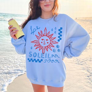 May include: Light blue sweatshirt with a red and blue graphic design. The design includes a sun with a face, the word "SOLEIL", and other artistic elements. A yellow insulated beverage holder is held in the hand.