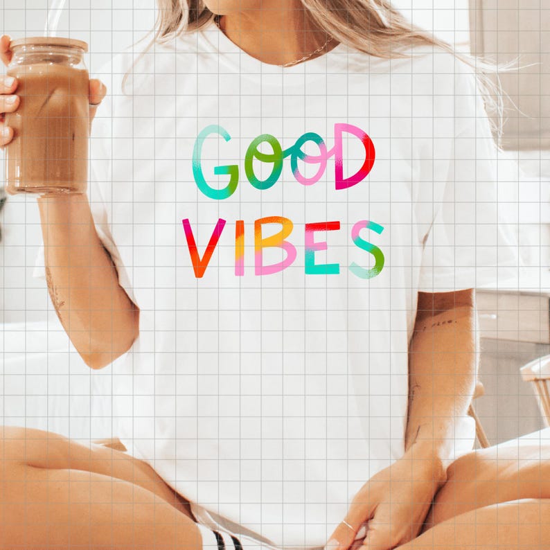 May include: White t-shirt with the words "Good Vibes" in colorful, gradient lettering. The shirt is being held by a person, and they are also holding a glass jar with a straw and a brown liquid inside.