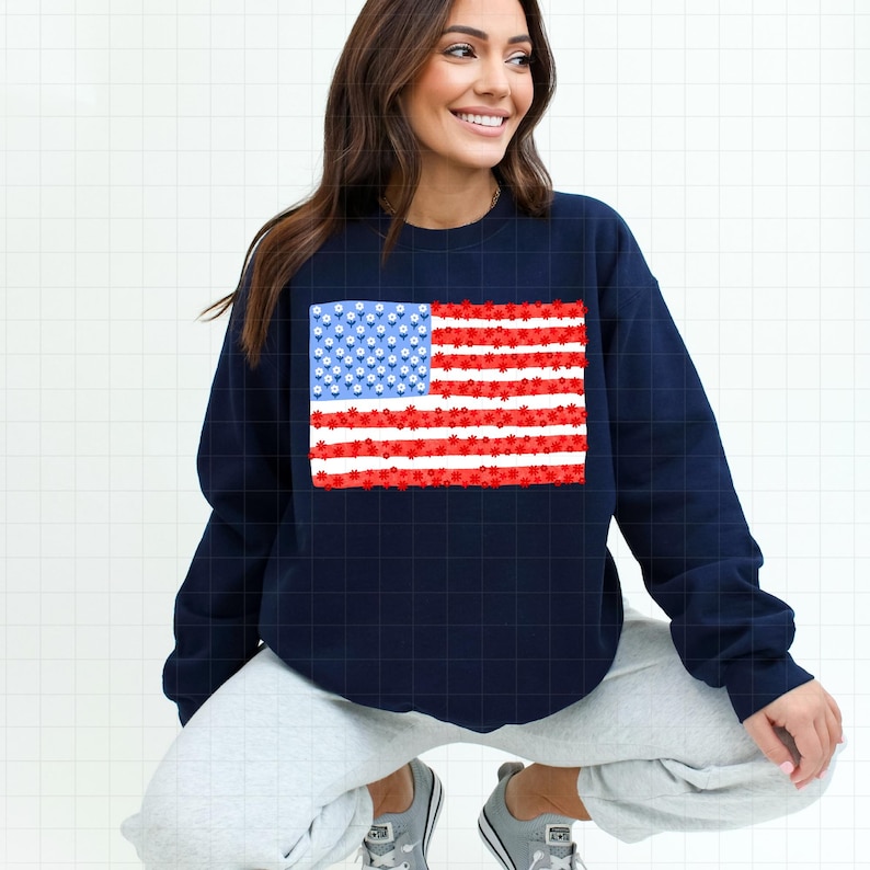 May include: Navy blue sweatshirt featuring a floral American flag design. The flag's stars are represented by small white and blue flowers, and the stripes are red flowers on white stripes. The sweatshirt is a casual, patriotic apparel item.