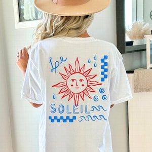 May include: White t-shirt with a red and blue graphic design. The design features a red sun with a face, blue raindrops, and the word "SOLEIL" in blue. The word "be" is written in blue script above the sun.