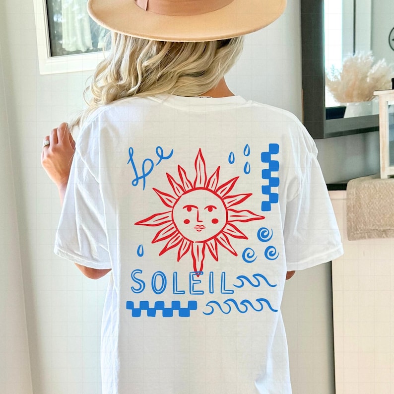 May include: White t-shirt with a red and blue graphic design. The design features a red sun with a face, blue raindrops, and the word "SOLEIL" in blue. The word "be" is written in blue script above the sun.