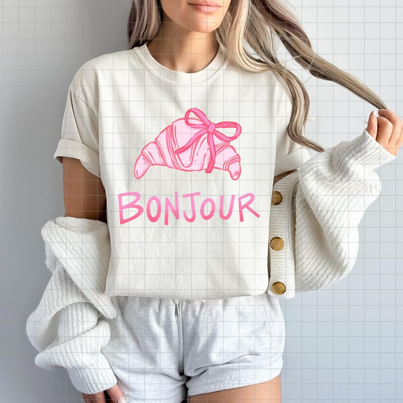 Op de afbeelding: Cr&egrave;mekleurig T-shirt met een roze croissant met strik en het woord "Bonjour" in het roze. Het shirt heeft korte mouwen en een relaxte pasvorm. Het ontwerp heeft een casual en vrouwelijke uitstraling.