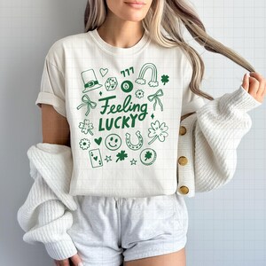 May include: Cream-colored t-shirt with a green graphic design featuring a leprechaun hat, dice, a rainbow, and the words "Feeling Lucky." The design includes a horseshoe, four-leaf clovers, and playing cards.