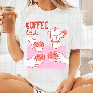 May include: White t-shirt with a pink graphic featuring coffee-related illustrations and the words "COFFEE Club" in red. The graphic includes a coffee maker, hands holding cups of coffee, and latte art.