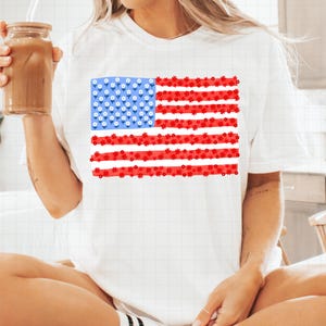 May include: White t-shirt featuring a floral American flag design. The flag's stars are represented by small white flowers on a blue background, and the stripes are red flowers. A casual and patriotic apparel item.
