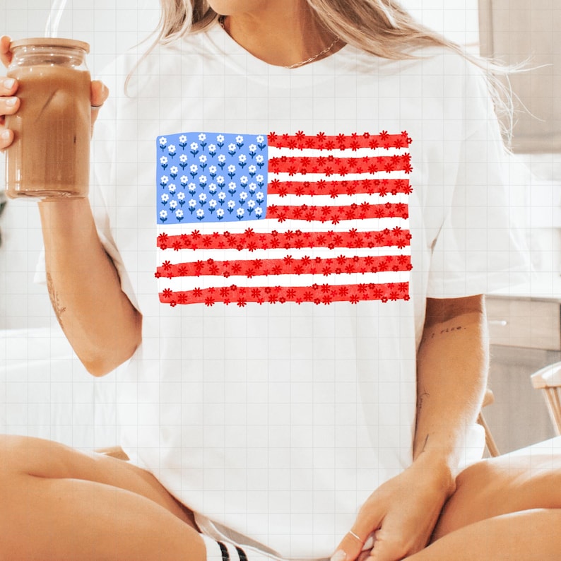 May include: White t-shirt featuring a floral American flag design. The flag's stars are represented by small white flowers on a blue background, and the stripes are red flowers. A casual and patriotic apparel item.