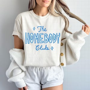 May include: Cream-colored t-shirt with the text "The Homebody Club" in blue script. The shirt has a crew neck and short sleeves. The model is wearing gray shorts and a white cardigan.