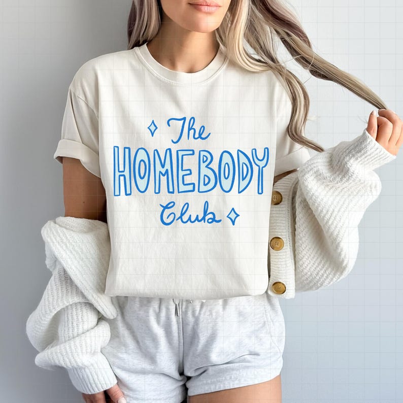 May include: Cream-colored t-shirt with the text "The Homebody Club" in blue script. The shirt has a crew neck and short sleeves. The model is wearing gray shorts and a white cardigan.