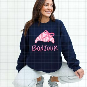 Op de afbeelding: Marineblauw sweatshirt met een roze croissantontwerp, versierd met een strik. Het woord "Bonjour" staat in roze onder de croissant. Het sweatshirt heeft een casual stijl.