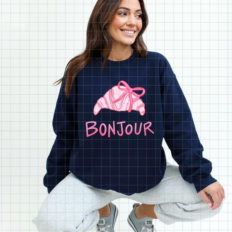 Op de afbeelding: Marineblauw sweatshirt met een roze croissantontwerp, versierd met een strik. Het woord "Bonjour" staat in roze onder de croissant. Het sweatshirt heeft een casual stijl.