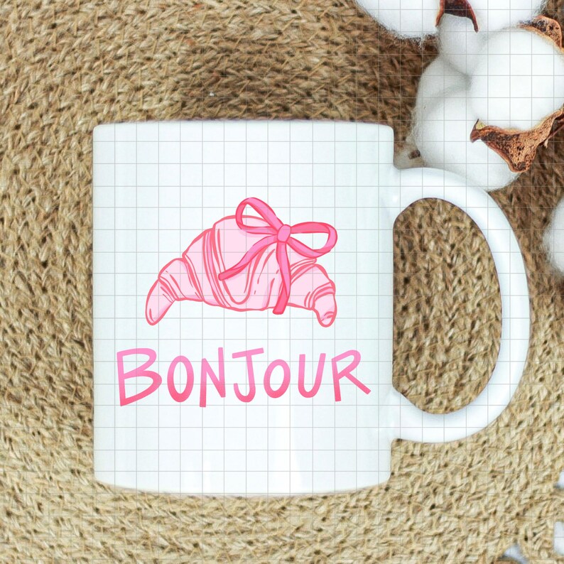 Op de afbeelding: Witte keramische mok met een roze croissant illustratie vastgebonden met een strik, en het woord "BONJOUR" in het roze. De mok heeft een gebogen handvat. De achtergrond is getextureerd.