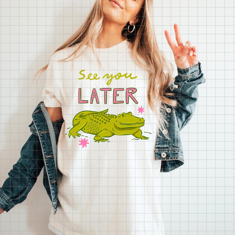 May include: Cream-colored t-shirt featuring a cartoon alligator in shades of green. The text "See you LATER" is above the alligator in green and pink. Two pink starbursts are on either side of the alligator.