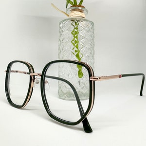 Peut inclure: Lunettes carrées noires et or rose avec des verres transparents. Les lunettes sont posées sur une surface blanche devant un vase en verre transparent avec un arrangement de fleurs blanches.
