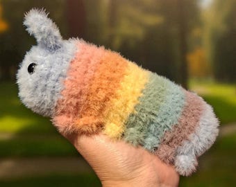 May include: A plush, rainbow-colored caterpillar toy with a gray head and tail. The caterpillar is made of soft, fuzzy material and features stripes of pink, orange, yellow, green, and brown. The toy is held in a person's hand.