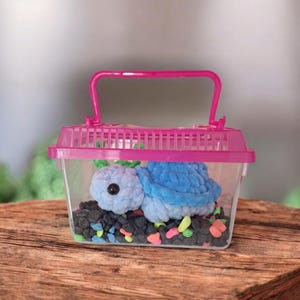 Puede incluir: Un portador de plástico rosa con un contenedor de plástico transparente en el interior. El contenedor contiene un juguete de peluche de tortuga azul y morado y piedras de colores.