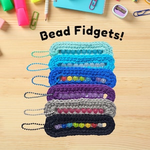 Peut inclure: Une pile de six fidgets à perles crochetés, de couleurs variées : bleu clair, bleu foncé, violet, gris et noir. Chaque fidget comporte une rangée de perles colorées et une boucle de chaîne. Le texte "Bead Fidgets!" est affiché au-dessus.