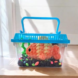 以下が含まれることがあります： 青い取っ手付きの小さなプラスチック製の魚タンク。タンクの中には、オレンジと黄色の毛糸で編まれた魚、緑色のプラスチック製の植物、黒と緑の石が入っています。
