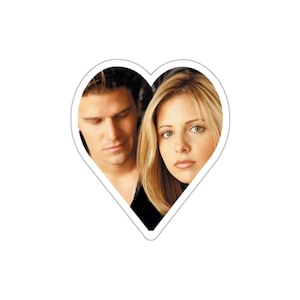 May include: A heart-shaped sticker featuring a close-up portrait of a couple. The man has dark hair and is wearing a black shirt. The woman has long blonde hair and is wearing a black shirt. They are looking at each other.