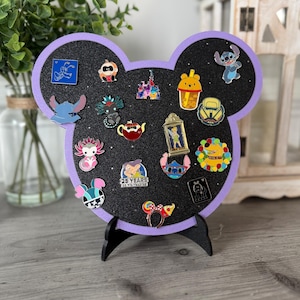 May include: A black Mickey Mouse-shaped pin display board with a purple border, holding various enamel pins. The pins feature cartoon characters and themed designs. The board is on a black stand.