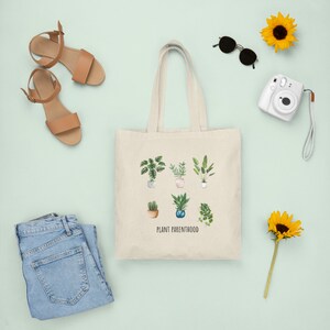 May include: A white canvas tote bag with the text "Plant Parenthood" and illustrations of potted plants. The bag is surrounded by various items, including sunglasses, a camera, sandals, and sunflowers.