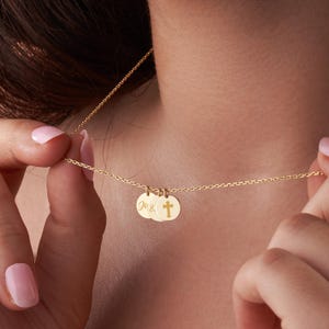 May include: A delicate gold necklace featuring three circular pendants. The pendants include engraved initials and a cross symbol. The necklace is displayed against a neutral background, highlighting the jewelry's design and craftsmanship.