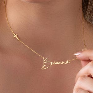 May include: A gold-colored necklace featuring the name "Brianna" in a cursive font. A small cross is on the left side of the name. The necklace is on a delicate chain.