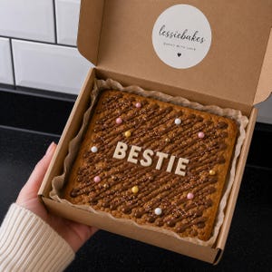 May include: A square cookie cake in a brown cardboard box with the word BESTIE in white icing letters. The cake is drizzled with chocolate and topped with colourful sprinkles and small round sweets. The box lid features a round lessiebakes logo.