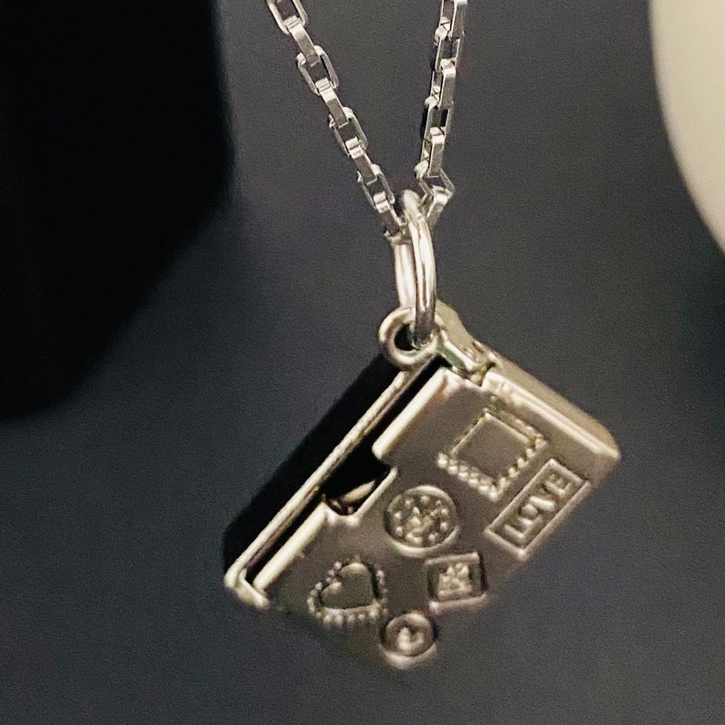 May include: Silver charm necklace featuring a small, intricate charm shaped like a closed book. The charm is engraved with a heart, stars, and the word "LOVE".