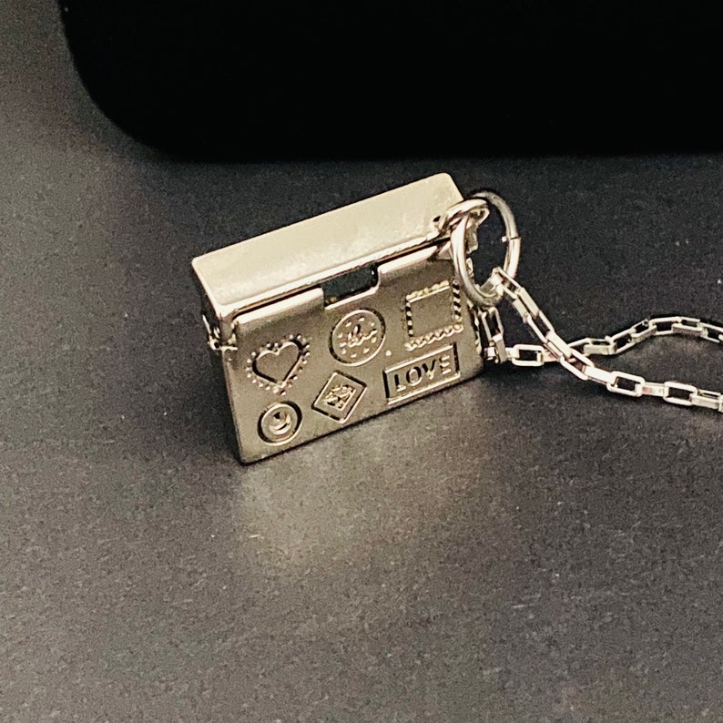 May include: A silver charm necklace with a small rectangular charm that opens. The charm has various symbols engraved on it, including a heart, a smiley face, a star, a square, and the word "LOVE".