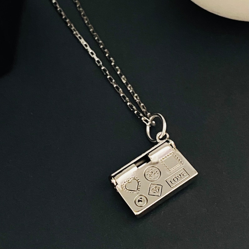 May include: A silver necklace with a small rectangular locket pendant. The locket has a heart, a circle, a diamond, and a square with the word "LOVE" engraved on it.