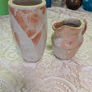 Może przedstawiać: Wysoki, cylindryczny wazon ceramiczny oraz mniejszy, zaokrąglony dzbanek ceramiczny, oba z nakrapianą, kremową glazurą bazową, teksturowanymi, czerwono-pomarańczowymi abstrakcyjnymi plamami oraz szorstkim, ręcznie wykonanym wykończeniem.