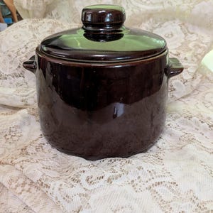 May include: A dark brown ceramic bean pot with a matching lid and two small side handles. The pot features a smooth, glossy finish and a cylindrical body shape. The lid has a central knob handle, and the overall design is simple and functional.